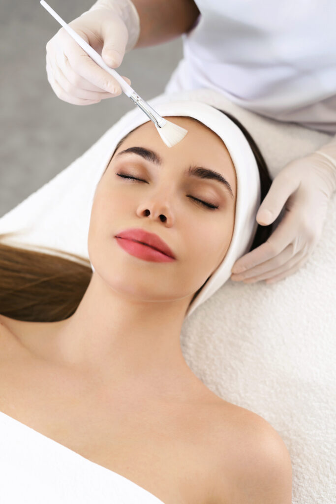 Model in white headband bandage cap lying on couch with closed eyes. Hand of beautician doctor in white glove touching her face with brush. Head and shoulders, healthcare, cosmetology, beauty clinic